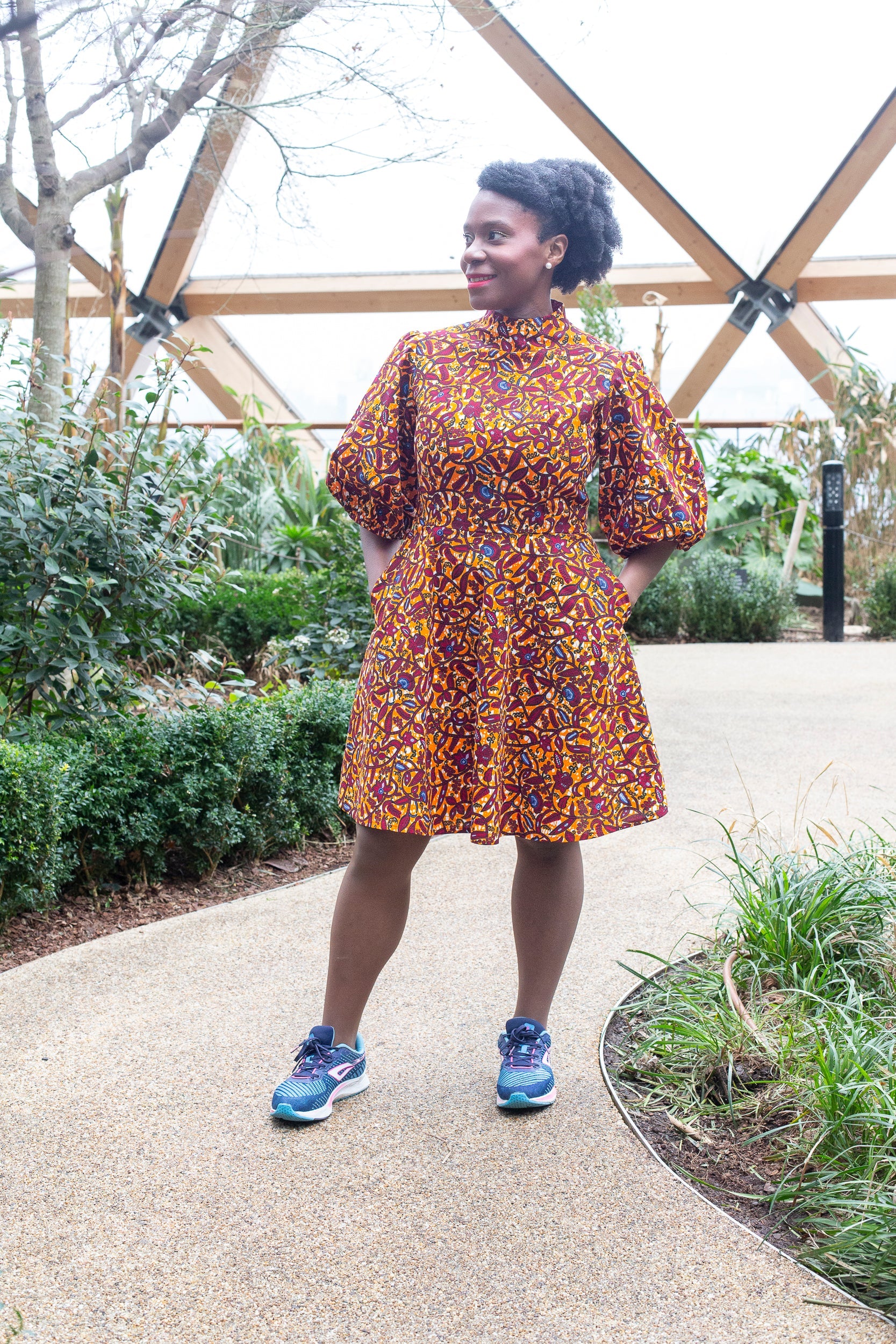 A person exudes happiness in an orange puff-sleeve dress, paired with blue trainers in a garden setting.