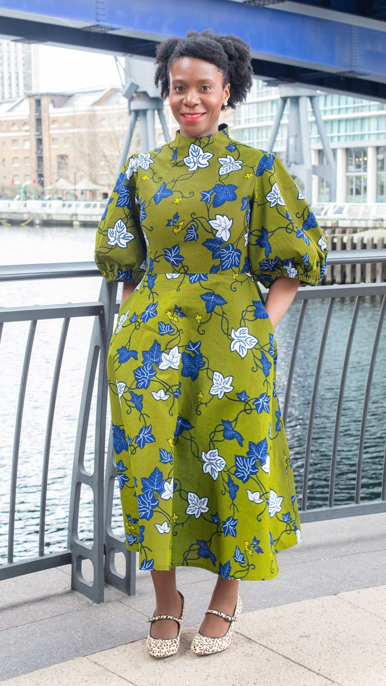 A woman stands in a khaki dress adorned with detailed blue and white leaves, paired with mary jane heels.