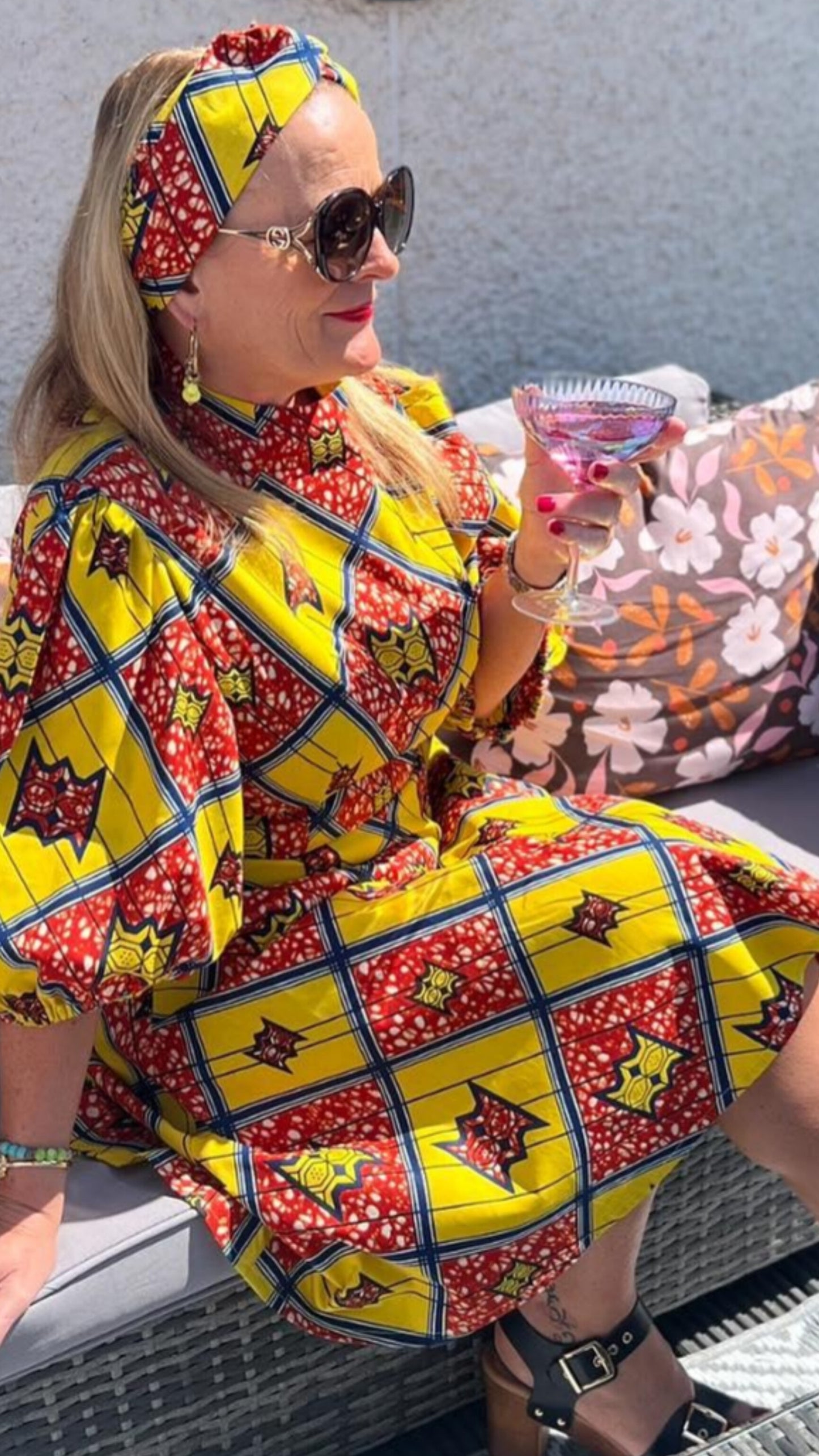A woman sits comfortably on a couch, holding a drink, and exudes a joyful vibe in a yellow and red short puff-sleeve dress with a matching print hairband. Wearing sunglasses, they radiate a carefree and stylish demeanour.