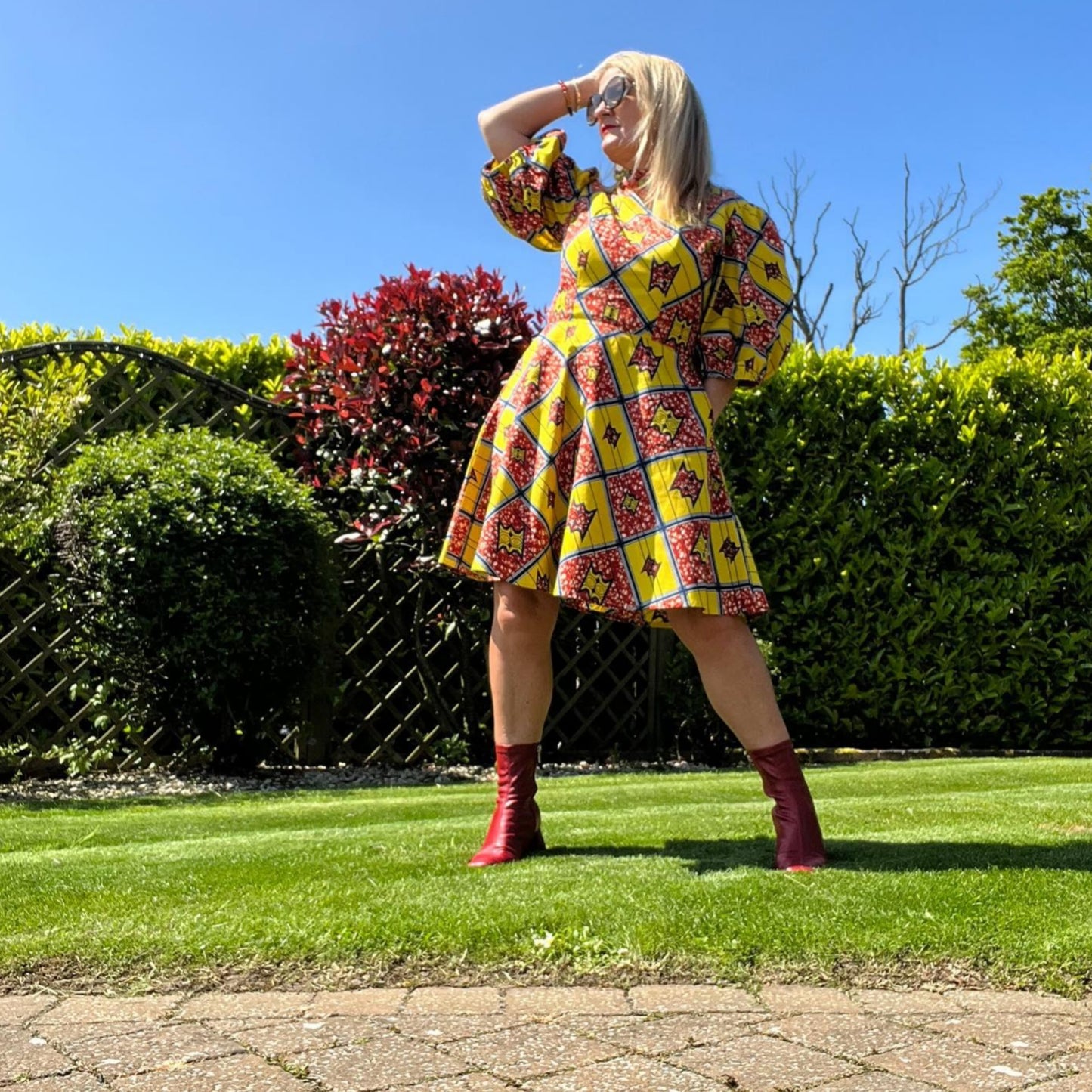 A woman confidently posing in front of a garden setting, wearing the yellow red puff sleeve short dress, paired with red boots and sunglasses.