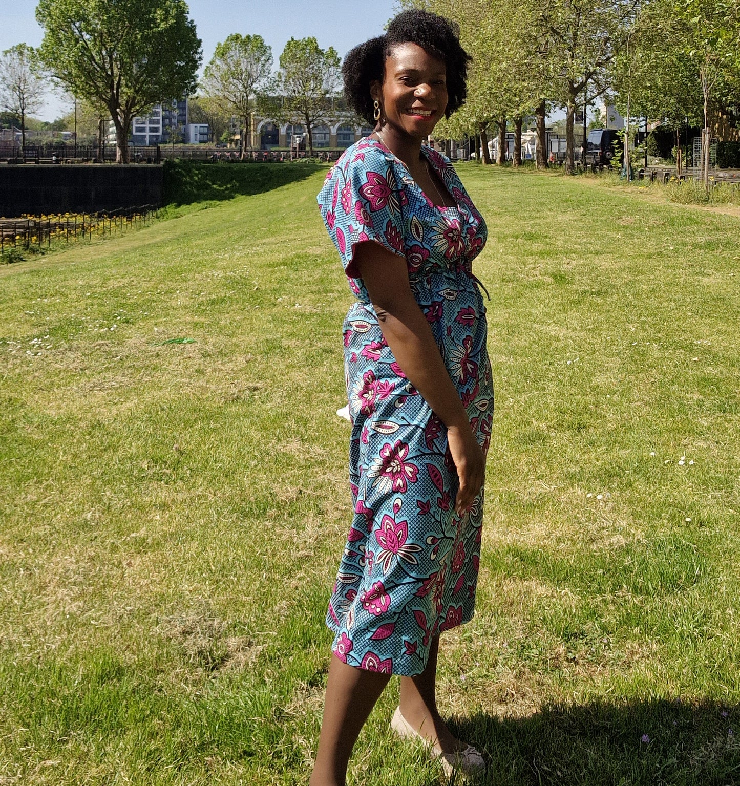 In a park setting, a woman confidently poses wearing a chic blue kaftan dress embellished with a delightful pink floral print. 