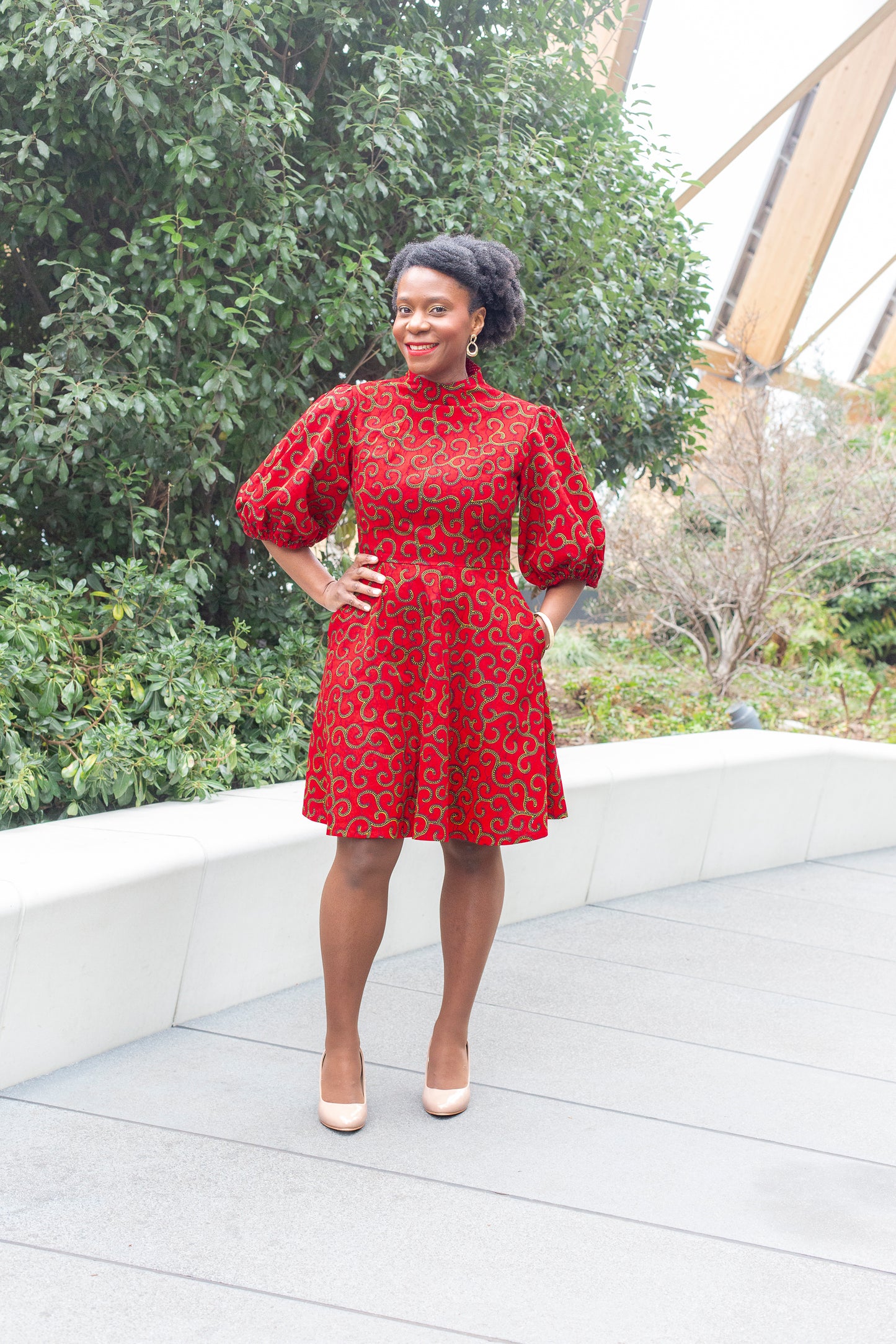 A woman confidently posing in a red puff sleeve dress paired with beige heels in a park setting.