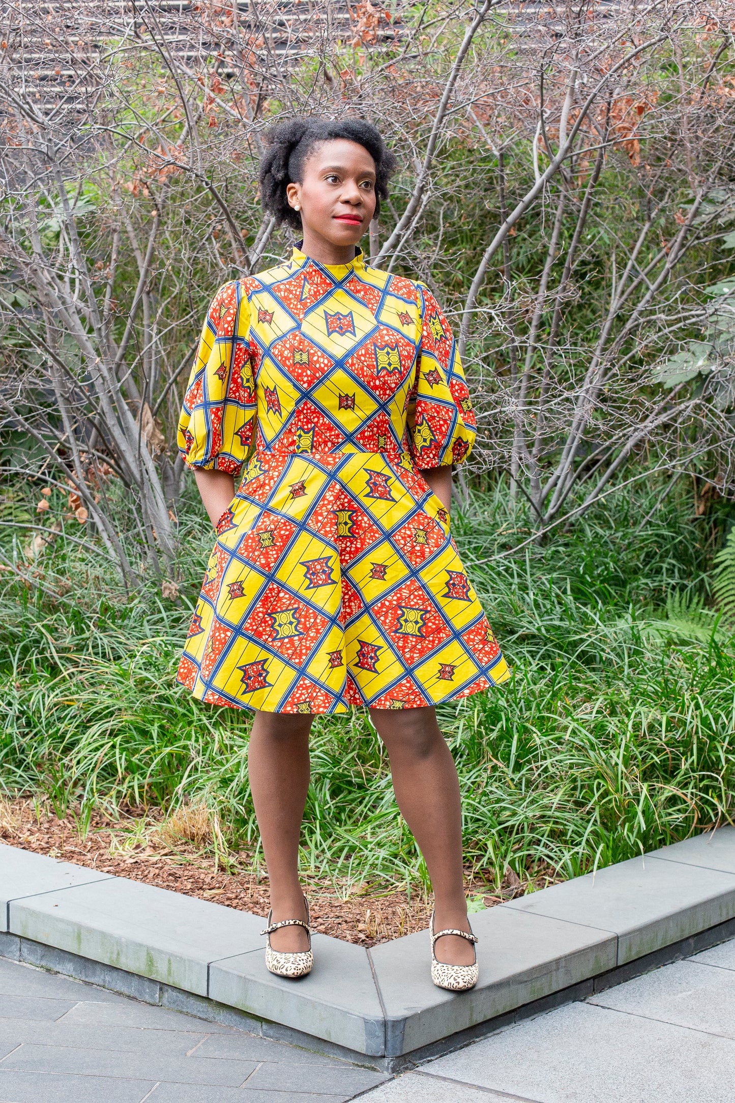 A woman in a short yellow puff-sleeve dress with red elements, paired with white Mary Jane flats, posing in front of a garden setting.