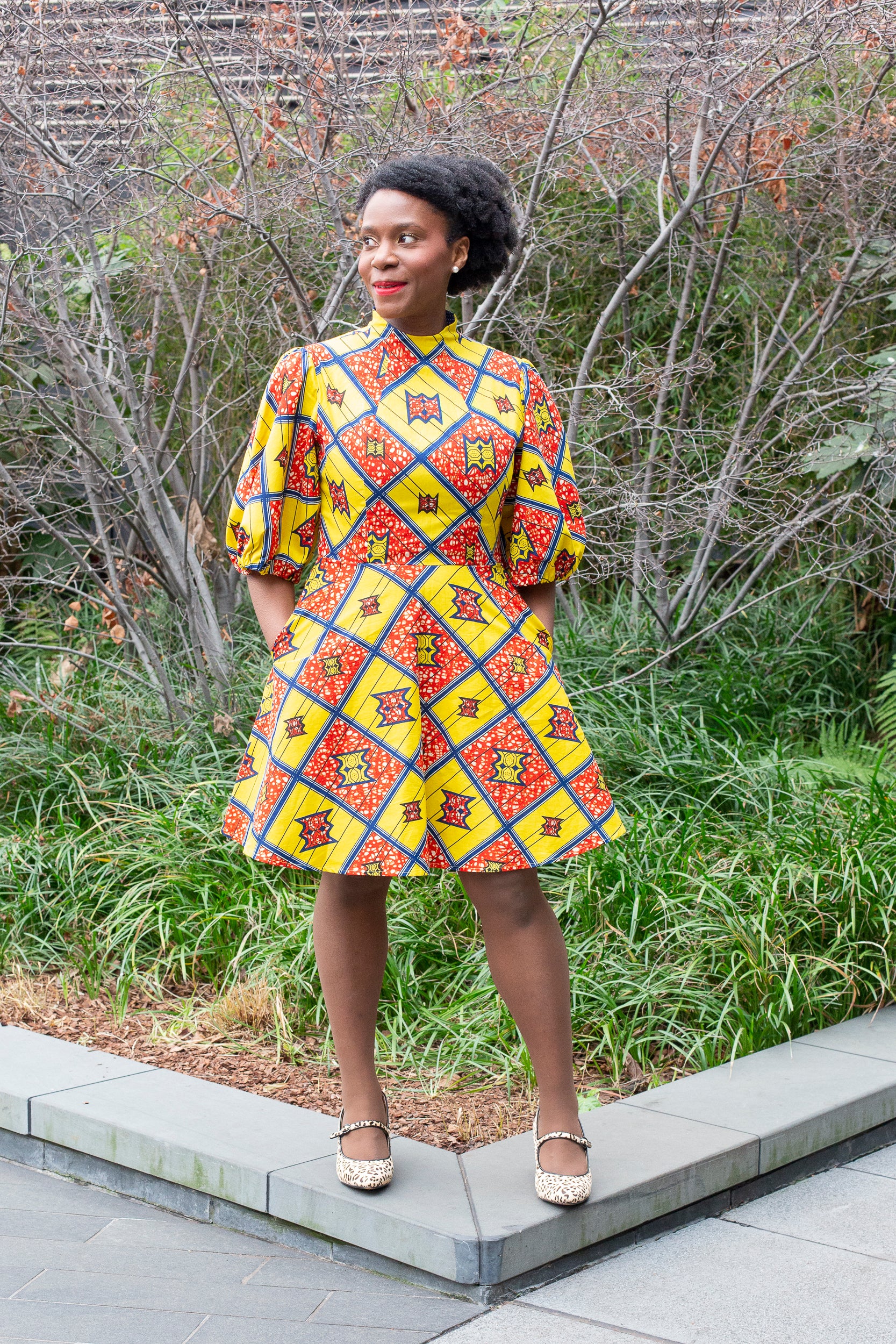 A person radiates happiness in a short yellow and red puff-sleeve dress, paired with white Mary Jane flats, standing in front of a garden setting.