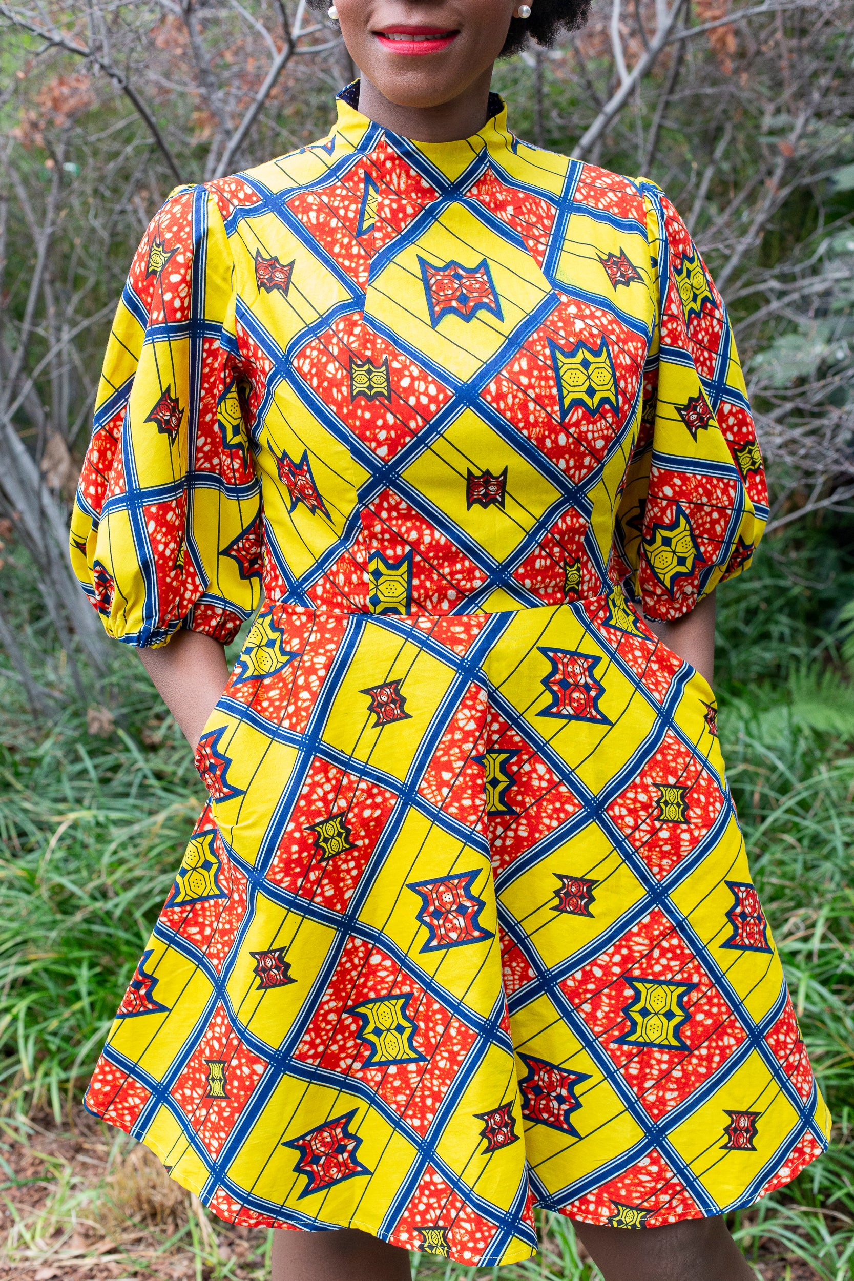 a close up of a woman wearing the short puff sleeve yellow and red print dress, who places hands in her pockets to showcase the comfortability of the dress.
